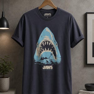 May include: A dark grey short-sleeve t-shirt featuring a graphic of a large shark with an open mouth emerging from water. The text JAWS and UNIVERSAL STUDIOS appears at the neckline, with JAWS 50TH ANNIVERSARY printed below the shark graphic.