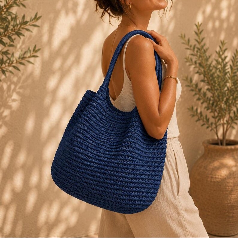 May include: A deep navy blue crochet tote bag featuring a textured, ribbed knit pattern with two integrated shoulder straps. The bag has a spacious, open-top design and is constructed from a thick, durable yarn, showcasing a consistent stitch.