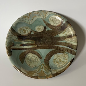 Op de afbeelding: Een ronde keramische schaal met een licht saliegroene basisglazuur, donkerbruine, met een penseel aangebrachte abstracte patronen en cirkelvormige, getextureerde crèmekleurige accenten die in een organische opstelling over het oppervlak zijn verdeeld.