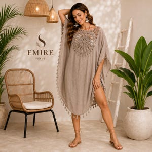 May include: A beige kaftan dress featuring a large circular crochet panel on the chest, short sleeves, and a hemline adorned with small hanging tassels. The garment has a loose, flowing silhouette and is made of a lightweight, textured fabric.
