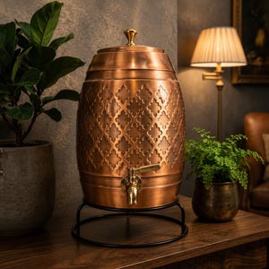 Puede incluir: Un dispensador de bebidas de cobre con forma de barril que presenta un patrón de cuatrifolio en relieve, un grifo de latón pulido y una tapa a juego con un pequeño pomo. El dispensador descansa sobre un soporte circular de metal negro.