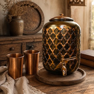 Puede incluir: Un dispensador de bebidas cilíndrico de metal con acabado en bronce oscuro y un patrón de recortes de cuatrifolio, con tapa a juego y grifo metálico. Dos vasos de color cobre liso se encuentran junto al dispensador sobre una base de madera.