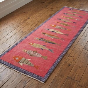 May include: A rectangular runner rug in a vibrant pink hue featuring a repeating pattern of stylised fish in various colours including orange, blue, and beige. The rug is framed by a dark blue border with a subtle geometric pattern and short fringe.