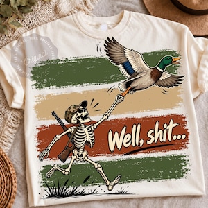 May include: A cream-colored t-shirt featuring a graphic of a skeleton wearing a camouflage cap and carrying a rifle. The skeleton is reaching up to grab a flying mallard duck. The text "Well, shit..." is printed in a bold, dark font.