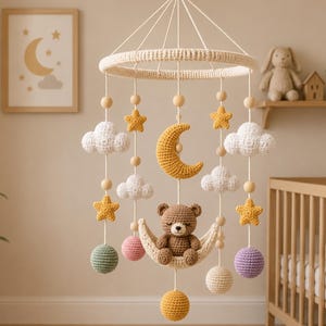 May include: A hanging nursery mobile featuring a central brown crocheted bear sitting on a crescent moon. The mobile includes white crocheted clouds, yellow stars, and multi-colored spheres suspended from a circular frame with wooden beads.
