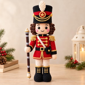 Puede incluir: Una muñeca cascanueces tejida a ganchillo con uniforme rojo y negro con detalles dorados, un sombrero negro alto con una pluma blanca y cabello castaño rizado. La muñeca sostiene un bastón con rayas marrones y blancas y tiene mejillas rosadas.