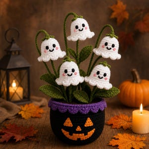 May include: A crochet plant featuring five white ghost-shaped flowers with black eyes and pink cheeks, each on a green stem with green leaves. The plant is potted in a black crochet container with an orange jack-o'-lantern face design.