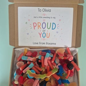 May include: A brown cardboard gift box filled with an assortment of colourful gummy sweets. The inside lid features a white label with the text: To Olivia, Just a little something to say PROUD OF YOU, Love From Roxanne, decorated with small hearts.