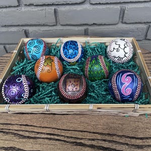 Puede incluir: Siete pelotas de béisbol decoradas con diseños pintados a mano en azul, naranja, morado, verde y blanco, con patrones de espirales, escamas y motivos florales, dispuestas en una cesta rectangular de bambú con hierba de papel verde.
