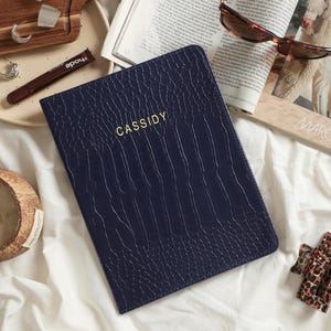 May include: A dark blue notebook with a textured crocodile-skin pattern and the name CASSIDY embossed in gold lettering on the front cover. The notebook is positioned on a white fabric surface alongside a magazine, sunglasses, and jewelry.