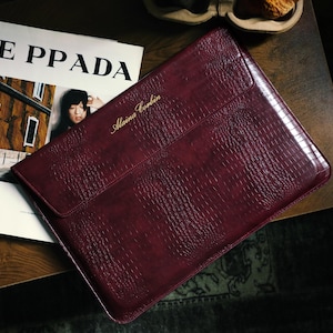 May include: A deep burgundy laptop sleeve with a textured crocodile-skin pattern and the gold-embossed name Alaina Corbin on the front flap. The sleeve is positioned next to a magazine with the visible text KE PPADA on the cover.