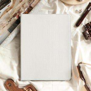 May include: A rectangular white tablet case featuring a textured crocodile skin pattern. The case has a clean, minimalist design with visible stitching along the edges, providing a structured and protective cover for a digital device.