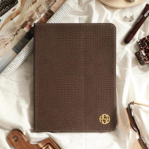 May include: A rectangular brown tablet case with a textured crocodile skin pattern and a gold circular monogram featuring the letters HSB in the bottom right corner. The case has visible stitching along the edges and a slim, flat profile.