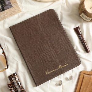 May include: A rectangular brown notebook with a textured crocodile-skin pattern and the name Evonne Marden embossed in gold script on the bottom right corner. The notebook is surrounded by various personal accessories including sunglasses and a tube.
