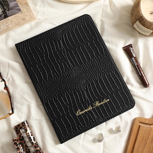 May include: A black rectangular notebook with a textured crocodile skin pattern and the name Querida Benton embossed in gold lettering on the front cover. The notebook is positioned centrally, surrounded by various personal accessories.
