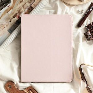 May include: A rectangular, light pink tablet case with a smooth, solid-coloured exterior and rounded corners. The case is positioned vertically, featuring a clean, minimalist design with visible stitching along the edges of the front cover.