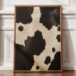 Peut inclure: Une œuvre d'art murale rectangulaire présentant une impression haute résolution d'un motif de peau de vache avec des taches irrégulières brun foncé et crème. L'œuvre est encadrée dans un cadre en bois fin de ton moyen aux bords nets.