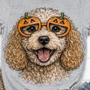 May include: A heather gray t-shirt featuring a graphic illustration of a curly-haired, light brown dog wearing orange pumpkin-shaped glasses. The dog has a happy expression with its mouth open and tongue visible, centered on the front of the shirt.