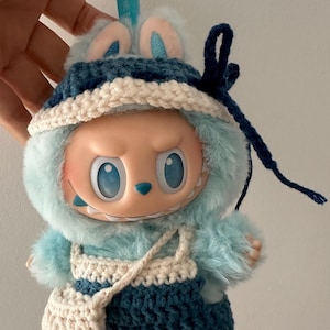 May include: A plush monster doll with light blue fur, wearing a dark blue crocheted hat with a white brim and matching dark blue crocheted overalls with a white strap and a small white side bag. The doll has large blue eyes and a light blue keychain.