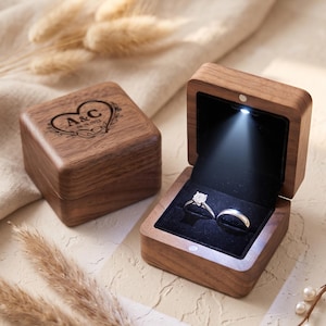 May include: Two square wooden ring boxes with a dark brown wood grain finish. One box is closed and features a laser-engraved heart design with the letters A&C and the date 09.28.2022. The other box is open, revealing a black velvet interior with an LED light illuminating a diamond engagement ring and a plain wedding band.