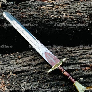 May include: A silver-toned sword featuring a double-edged blade with etched geometric lines. The ricasso displays a red shield emblem with a trident symbol and the Greek text ANAKAYMOΣ. It has a gold-colored crossguard and a brown leather-wrapped grip.