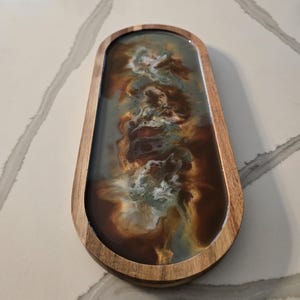 May include: An oval-shaped wooden tray featuring a smooth, polished finish. The center is filled with a poured resin inlay displaying a swirling, marbled pattern of deep brown, metallic gold, and soft blue-grey tones with white highlights.