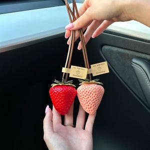 May include: Two strawberry-shaped charms with green leaves, one in bright red and one in light pink. Each charm hangs from a brown leather loop and features a small tan paper tag printed with the text "OH! PANCAKE" and "Strawberry" with a line drawing.