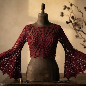 May include: A dark red long-sleeved top featuring an intricate crochet spiderweb pattern throughout the bodice and wide, flared bell sleeves. The garment is displayed on a vintage-style mannequin, showcasing the open-knit texture and design.