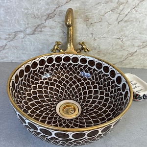 Puede incluir: Un lavabo de sobreponer redondo de cerámica con un patrón de escamas en marrón oscuro y blanco, borde dorado y desagüe a juego. Un grifo de latón texturizado de estilo antiguo está montado detrás del lavabo sobre una superficie plana.