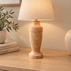May include: A table lamp featuring a light-coloured, natural wood base with a visible grain pattern and a tapered, cylindrical shape. The lamp is topped with a conical, off-white fabric lampshade that emits a soft, warm glow when illuminated.