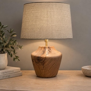 May include: A table lamp featuring a natural wood base with visible grain and knots, topped with a tapered, textured beige fabric lampshade. The lamp has a brass-coloured metal neck connecting the base to the shade, emitting a warm light.