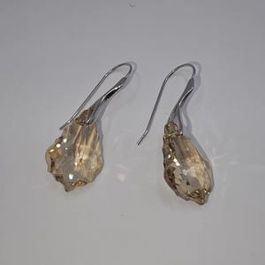 May include: A pair of drop earrings featuring faceted, champagne-coloured crystals suspended from silver-toned metal hooks. The crystals have an irregular, organic shape with multiple reflective surfaces that catch the light, creating a clear, sparkling appearance.