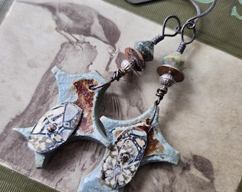 May include: A pair of handmade ceramic star earrings with a blue and white glaze. The earrings are accented with copper and silver beads, and green gemstone beads. The earrings are hanging from silver wire.