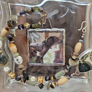 Può includere: Una collana artigianale con un ciondolo con un'illustrazione di un cane, completata da una catena di perline. La collana incorpora varie perline di pietra naturale nei toni del verde, marrone e crema, con una chiusura argentata.