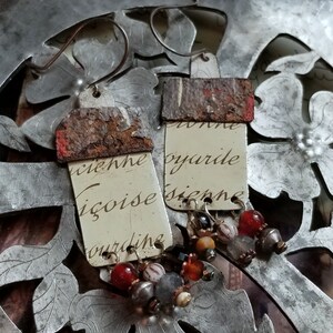 Pode incluir: Um par de brincos com uma estética rústica e vintage. Cada brinco apresenta uma peça retangular de metal com uma parte superior enferrujada e uma parte inferior de cor creme com texto escrito à mão. O texto no brinco esquerdo diz "wiehe igoise ourdine" e o texto no brinco direito diz "oyarde siepne". Cada brinco é adornado com um encanto pendente de contas em tons de vermelho, laranja, marrom e cinza.