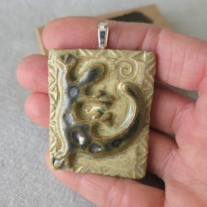 May include: A rectangular ceramic pendant featuring a lizard design. The pendant is mainly olive green with dark brown and grey accents. It has a silver-coloured bail for a necklace chain.