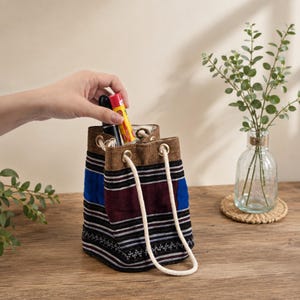 May include: A small drawstring bucket bag featuring horizontal stripes in shades of dark brown, maroon, and bright blue. The bag has a tan top band with metal grommets and a thick white rope drawstring. A yellow and red tube is inside the bag.