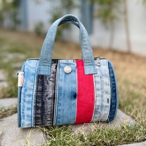 Puede incluir: Un bolso de mano con forma de barril construido con paneles verticales de mezclilla en varios tonos de azul claro, gris oscuro y una franja roja sólida. El bolso cuenta con dos asas de mezclilla, un detalle de botón de metal plateado y una etiqueta de tela blanca.