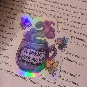 May include: A holographic sticker featuring a purple mug with the text 'a pixie pick me up' written on a banner. The mug emits swirling purple and blue steam, surrounded by three small fairies with wings, each holding a tiny star-tipped wand.