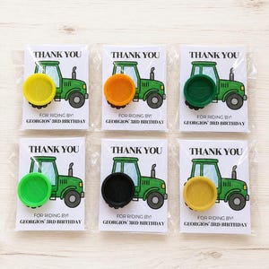 May include: Six party favor cards in clear plastic sleeves, each featuring a green tractor illustration and the text THANK YOU FOR RIDING BY! GEORGIOS' 3RD BIRTHDAY. Each card has a circular plastic wheel-shaped toy in yellow, orange, green, lime, black, or gold.
