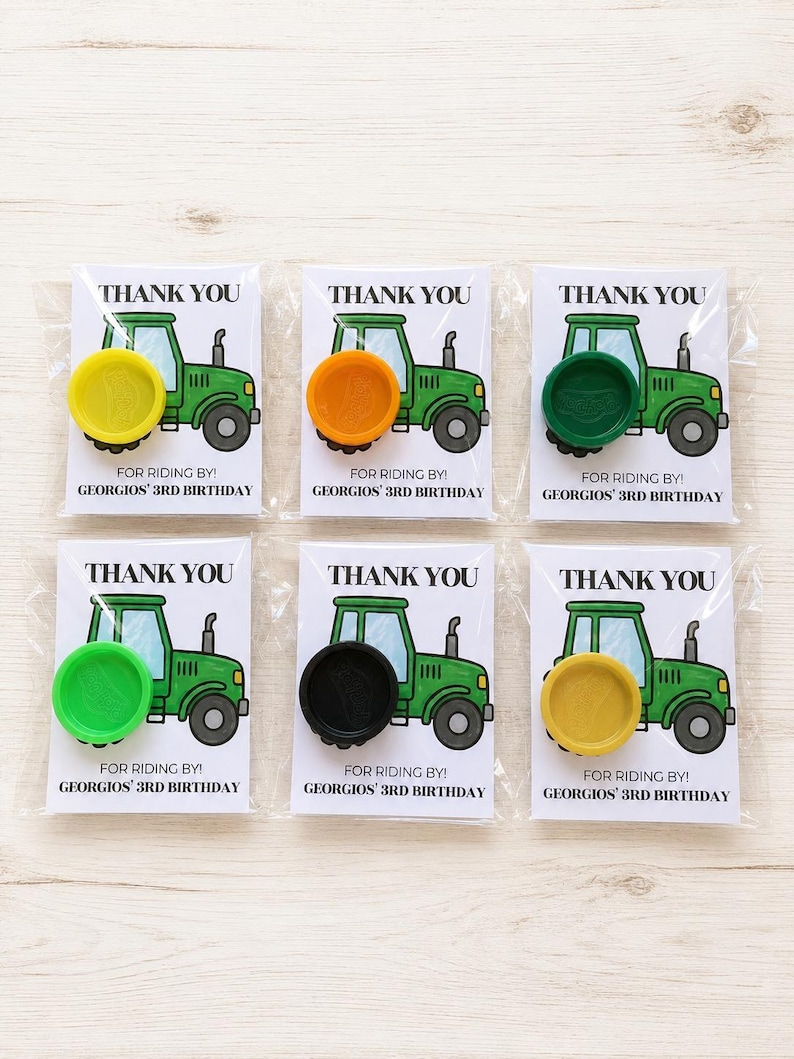 May include: Six party favor cards in clear plastic sleeves, each featuring a green tractor illustration and the text THANK YOU FOR RIDING BY! GEORGIOS' 3RD BIRTHDAY. Each card has a circular plastic wheel-shaped toy in yellow, orange, green, lime, black, or gold.