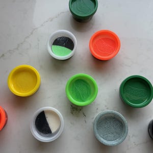 May include: A collection of nine circular plastic containers for modeling compound, featuring lids in various colors including green, orange, yellow, and black. Some containers are open, revealing multi-colored modeling compound inside.