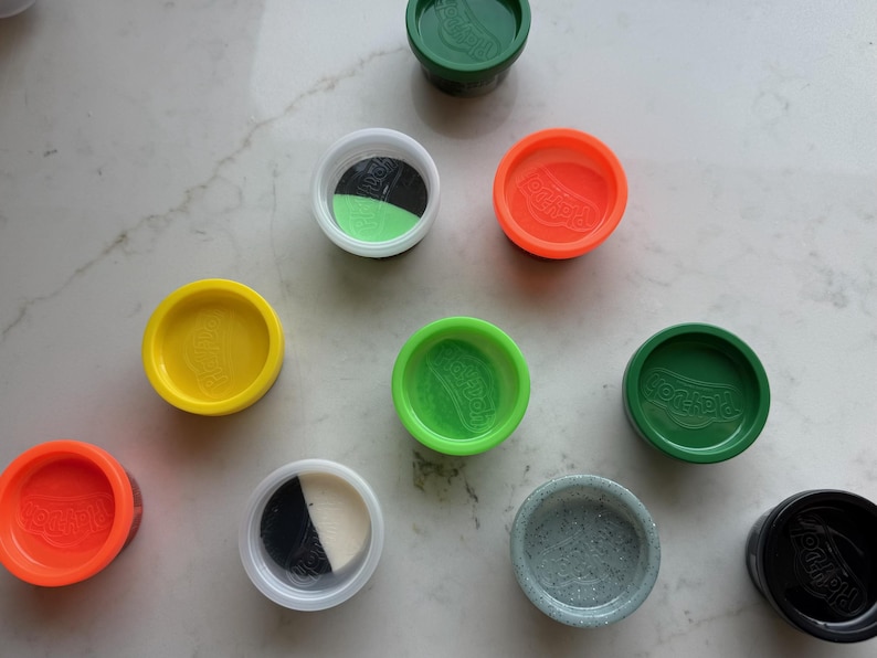 May include: A collection of nine circular plastic containers for modeling compound, featuring lids in various colors including green, orange, yellow, and black. Some containers are open, revealing multi-colored modeling compound inside.