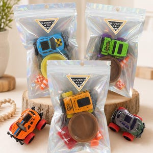May include: Three iridescent plastic pouches containing toy race cars, small circular plastic bases, and miniature orange traffic cones. Each pouch features a triangular sticker with the text OLIVER'S 4TH JAM. Two additional toy cars sit outside.