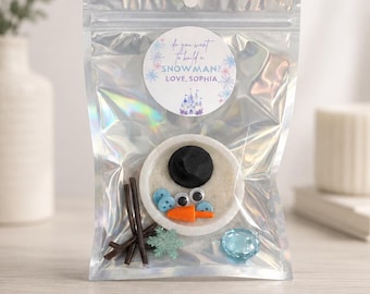 May include: A holographic pouch containing a snowman building kit with a white circular base, a black hat, googly eyes, blue buttons, an orange carrot nose, brown stick arms, and a blue snowflake. A sticker reads: Do you want to build a SNOWMAN? LOVE, SOPHIA.