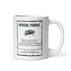 May include: A white ceramic mug featuring a black-bordered graphic with the text MOUCHE PERDUE and an illustration of a blue-green fly. The text includes French descriptions of the fly, ending with the email address sos.mouche.en.cavale@gmail.com.