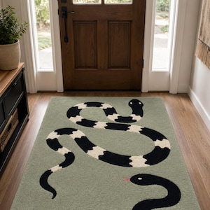 Puede incluir: Una alfombra rectangular de color verde salvia suave con dos serpientes. Una serpiente es larga con bandas alternas negras y blancas, mientras que la otra es una serpiente negra sólida con una pequeña lengua rosa visible cerca del borde.