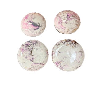 May include: Four circular plates featuring a marbled pattern in shades of white, soft pink, and dark purple. Each plate displays a unique swirling design with metallic gold accents integrated into the surface, creating a smooth, glossy finish.