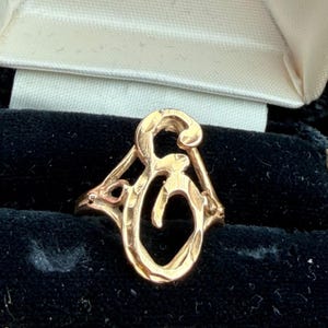 May include: A gold-toned ring featuring a stylized, open-work letter G design. The metal surface has a textured, hammered finish throughout the script-style character, which is integrated into a triangular frame that connects to the ring band.