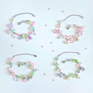 Peut inclure: Quatre bracelets à breloques composés de chaînes argentées et de perles colorées aux tons pastel rose, vert, bleu et jaune. Les bracelets présentent diverses formes comme des étoiles, des cœurs et des fleurs, avec des anneaux métalliques éparpillés.